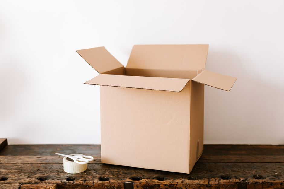 Caja de cartón abierta y vacía sobre una mesa de madera, junto a cinta adhesiva y unas tijeras