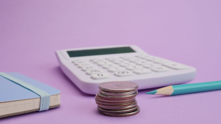 Calculadora, una pila de monedas y una libreta sobre una mesa lila